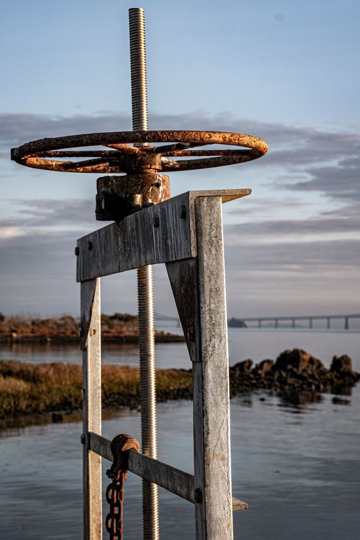 Rustic water pump with rusted wheel by a serene lakeside, bridge in the distance.
